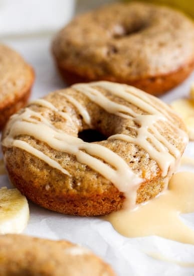 A close-up image of a glazed banana doughnut, with more doughnuts and banana slices in the background.