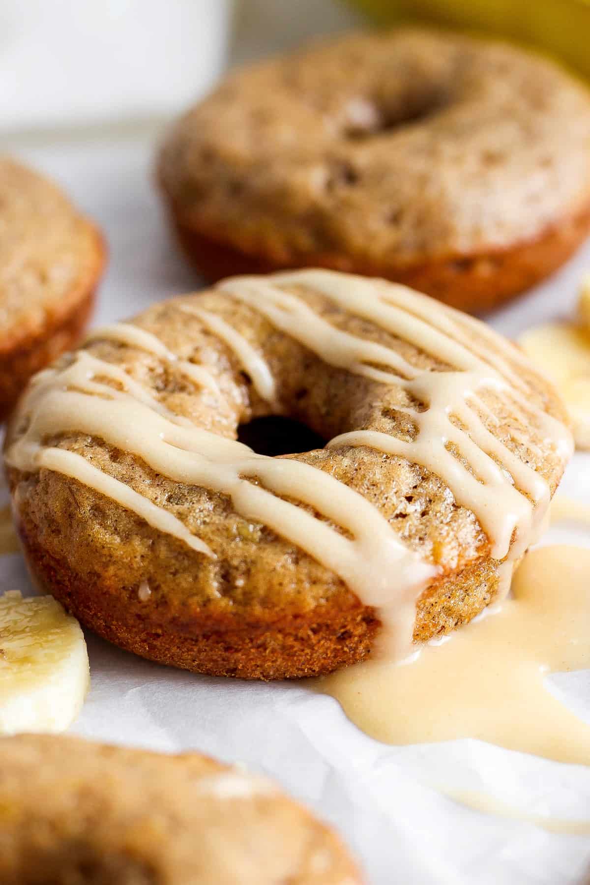 A close-up image of a glazed banana doughnut, with more doughnuts and banana slices in the background.