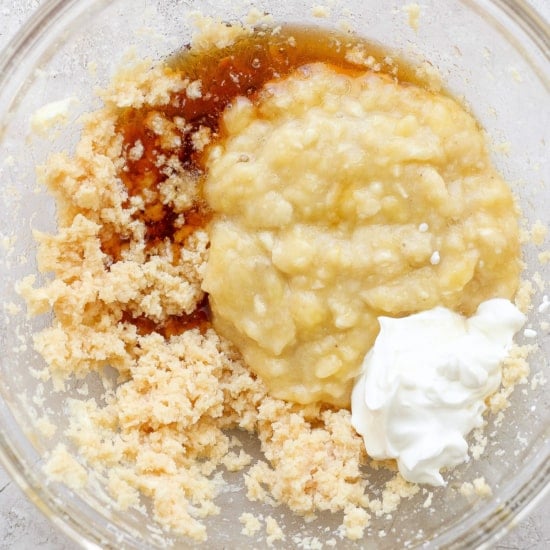 Ingredients combined in a glass bowl, possibly for baking, including mashed bananas, sugars, and a dollop of sour cream or yogurt.
