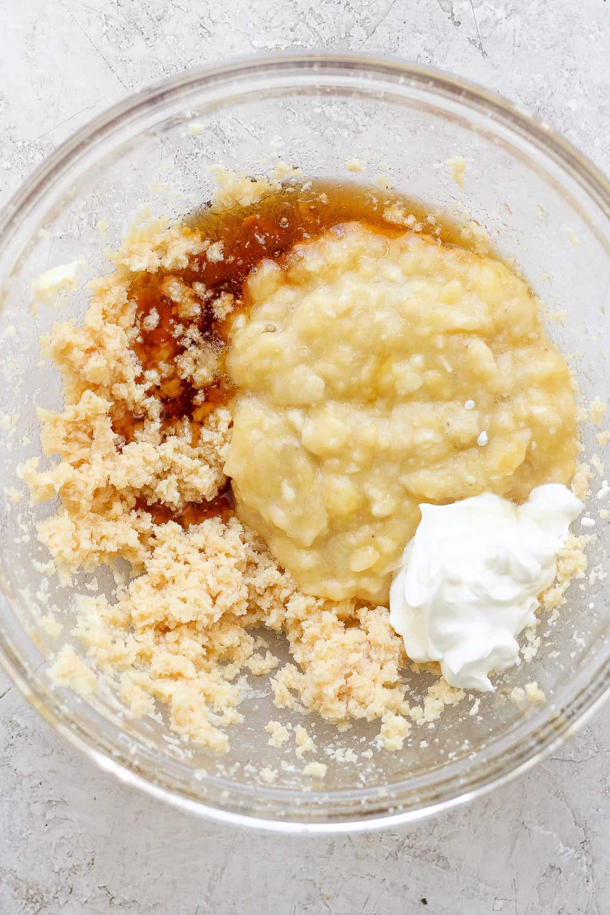 Ingredients combined in a glass bowl, possibly for baking, including mashed bananas, sugars, and a dollop of sour cream or yogurt.