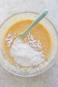 A glass bowl containing a mixture of flour and a liquid batter with a green silicone spatula.