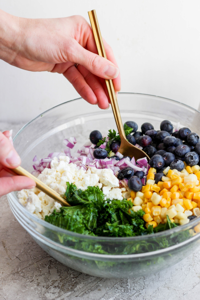 A person mixing a colorful salad with blueberries, corn, onions, and greens in a gl، bowl using a pair of golden tongs.