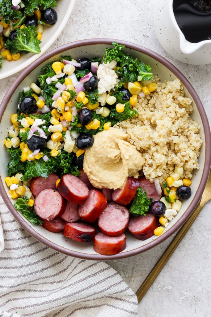 A bowl of quinoa, sliced sausage, and mixed vegetables, topped with a dollop of hummus.