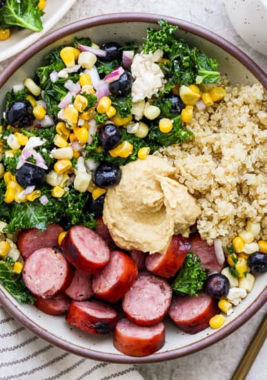 A bowl filled with quinoa, sliced sausage, hummus, kale salad, and a mix of corn and blueberries.