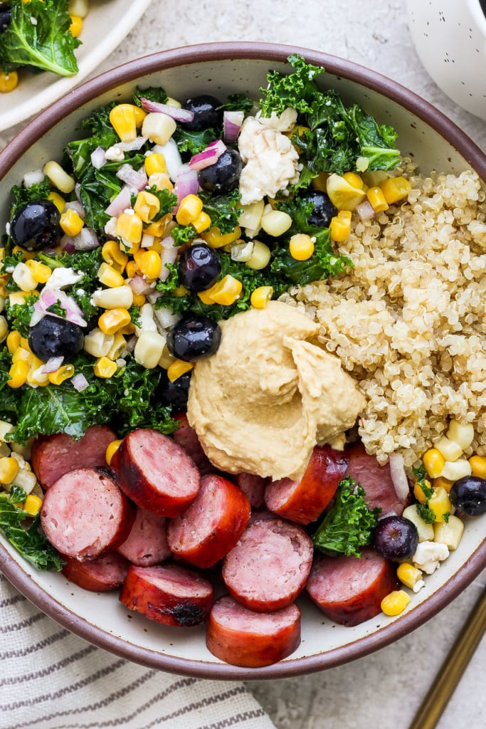 A bowl filled with quinoa, sliced sausage, hummus, kale salad, and a mix of corn and blueberries.