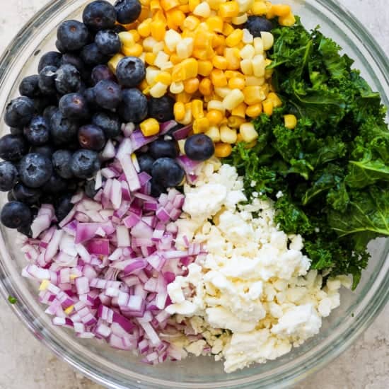 A bowl with a mix of ingredients including blueberries, corn, chopped red onions, kale, and feta cheese for a salad.