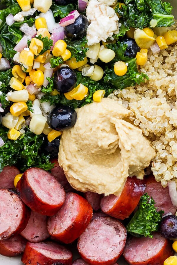 A bowl filled with quinoa, sliced sausage, kale, corn, black olives, onions, and a dollop of hummus.