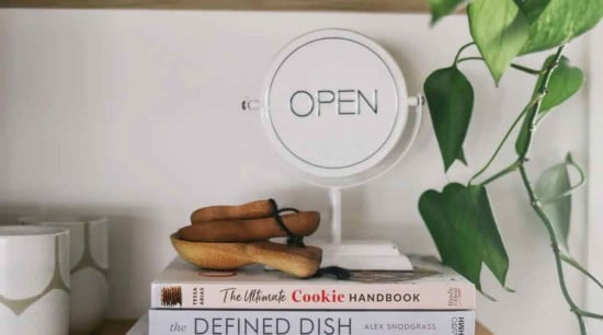 White "open" sign on a shelf above a stack of books with a pair of glasses and dried plant leaves.
