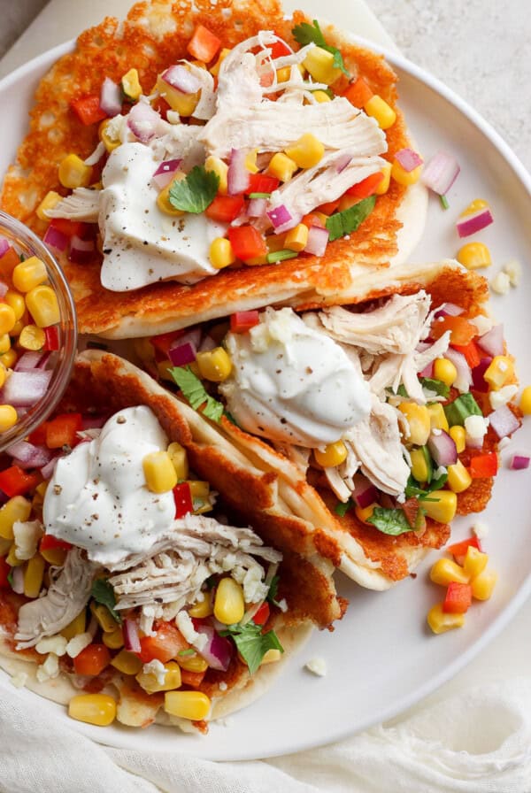 A plate containing chicken tacos garnished with corn, diced tomatoes, red onion, cilantro, and a dollop of sour cream, served on a white surface.
