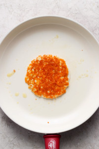 A single, fried hash brown resting on a white, circular non-stick pan with a red handle, displayed on a grey surface with a few scattered crumbs and oil droplets.