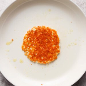 A single, fried hash brown resting on a white, circular non-stick pan with a red handle, displayed on a grey surface with a few scattered crumbs and oil droplets.