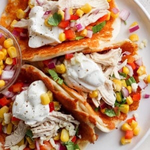 A plate of tacos with shredded chicken, corn, diced vegetables, and a dollop of sour cream served with a side of salsa.