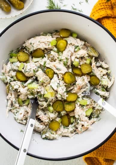 A bowl of dill pickle chicken salad with sliced pickles and fresh dill, accompanied by serving utensils.