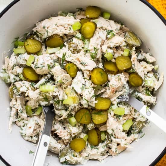 A bowl of dill pickle chicken salad with sliced pickles and fresh dill, accompanied by serving utensils.