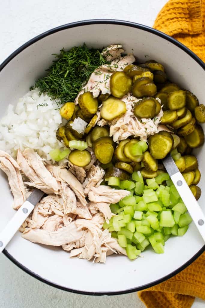 Ingredients for a recipe including shredded chicken, chopped onions, sliced pickles, and diced celery in a bowl with tongs.