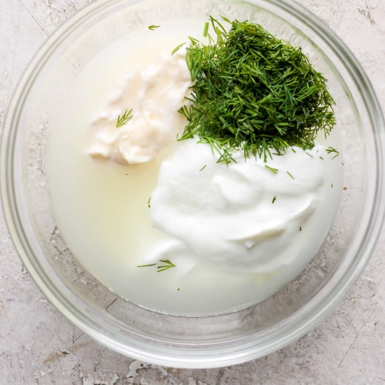 A glass bowl containing sour cream, dill, and a liquid, possibly part of a recipe preparation.