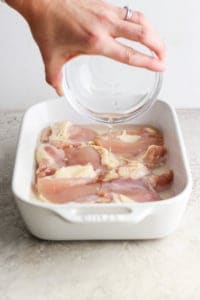 Pouring water over raw chicken in a baking dish.