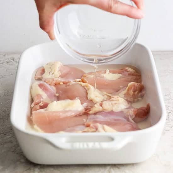 Pouring water over raw chicken in a baking dish.