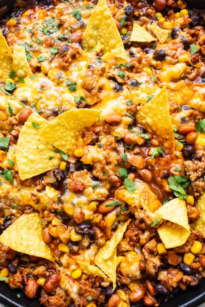 A skillet with nachos covered in melted cheese, ground meat, beans, corn, and garnished with fresh herbs.