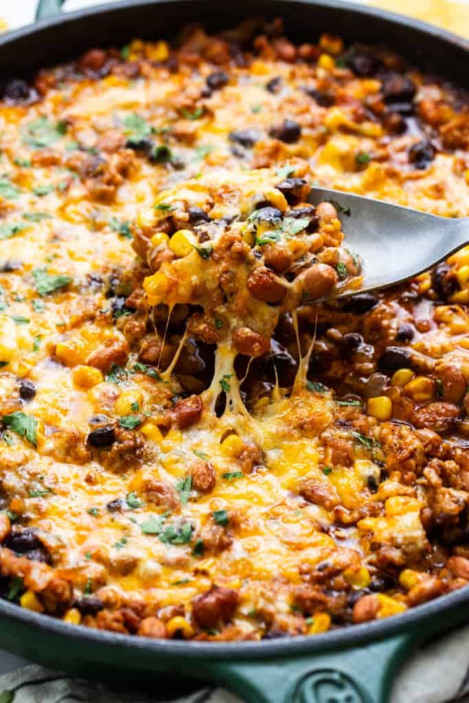 Skillet of baked cheesy chili with a spoon lifting a portion showing melted cheese stretch.