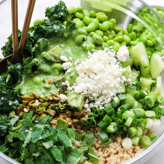 A vibrant bowl of healthy greens consisting of avocado, edamame, kale, and chopped herbs with a quinoa base, topped with crumbled cheese and served with a green dressing, ready to be.
