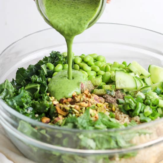 Pouring a green dressing over a bowl of salad with kale, quinoa, cucumber, and green peas.