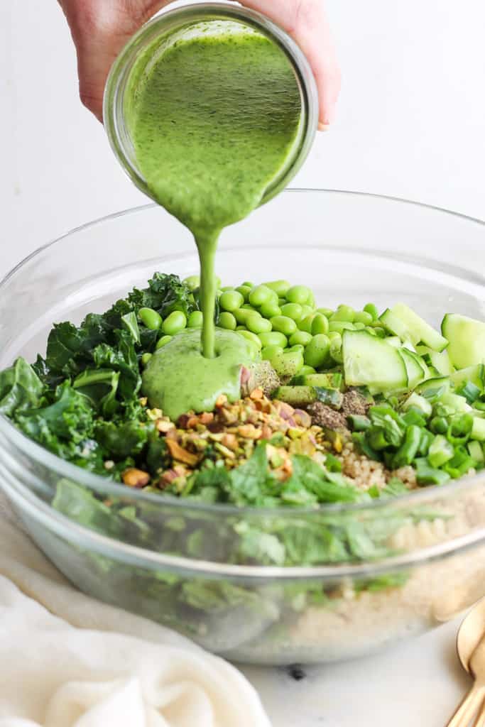 Pouring a green dressing over a bowl of salad with kale, quinoa, cucumber, and green peas.