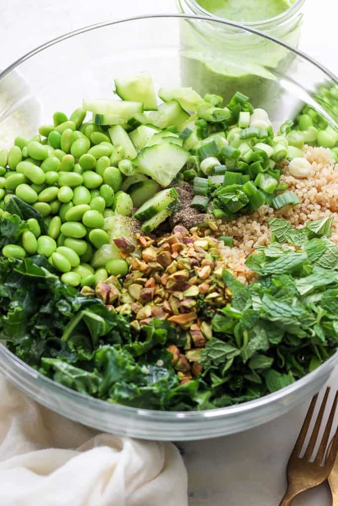 A bowl containing a variety of fresh ingredients including greens, grains, cucumber, peas, chopped nuts, and herbs, likely prepared for a healthy salad.