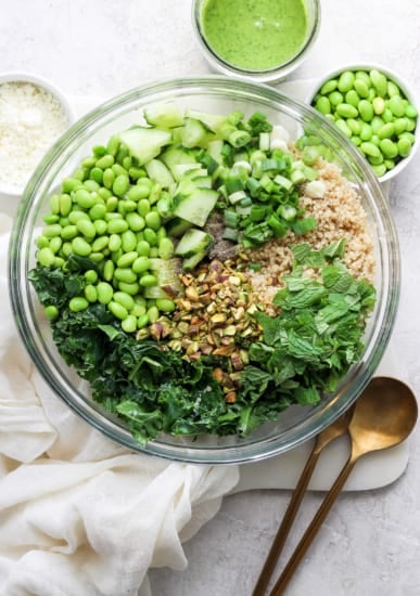 A bowl filled with various green ingredients including kale, cucumbers, peas, and chopped nuts, with a sprinkle of grated cheese, accompanied by a jar of green dressing and a spoon on a white.