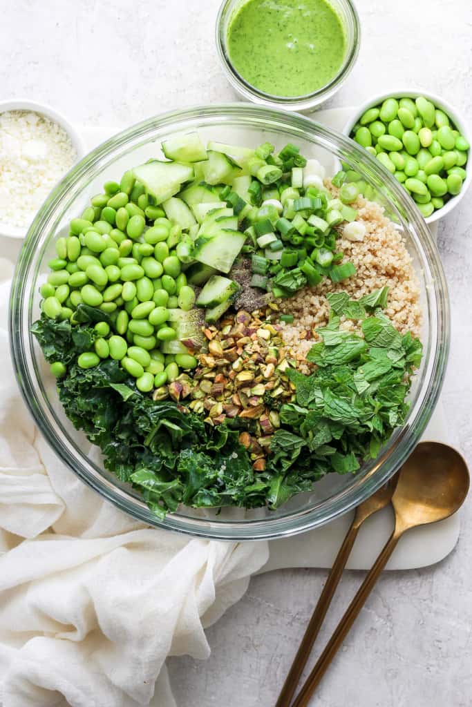 A bowl filled with various green ingredients including kale, cucumbers, peas, and chopped nuts, with a sprinkle of grated cheese, accompanied by a jar of green dressing and a spoon on a white.