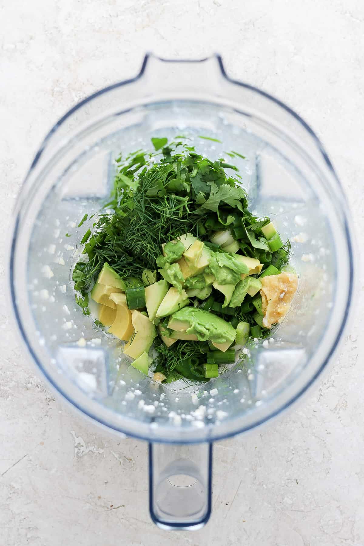 Ingredients including herbs, avocado, and banana prepared in a blender ready for blending.