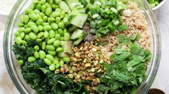 A bowl of salad containing quinoa, kale, chopped cucumbers, green onions, shelled edamame, chopped nuts, and fresh herbs, with a pair of golden spoons on the.