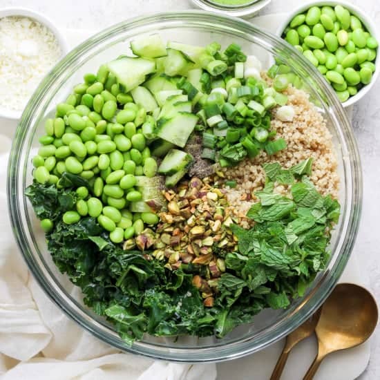 A bowl of salad containing quinoa, kale, chopped cucumbers, green onions, shelled edamame, chopped nuts, and fresh herbs, with a pair of golden spoons on the.