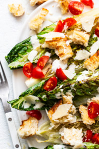 Grilled caesar salad with chicken, cherry tomatoes, croutons, and shaved parmesan cheese on a white plate.