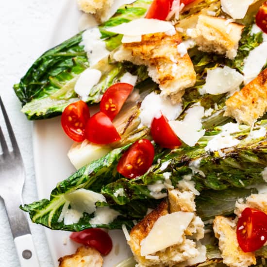 Grilled caesar salad with chicken, cherry tomatoes, croutons, and shaved parmesan cheese on a white plate.