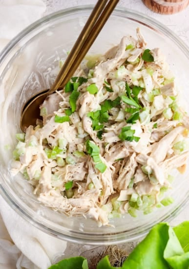 A bowl containing shredded chicken mixed with chopped celery and herbs, with a pair of gold-colored utensils.