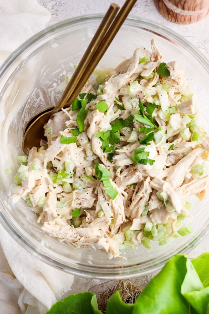 A bowl containing shredded chicken mixed with chopped celery and herbs, with a pair of gold-colored utensils.