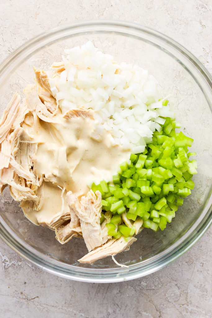 A glass bowl containing shredded chicken, diced onions, and chopped celery, possibly in preparation for a chicken salad or similar dish.