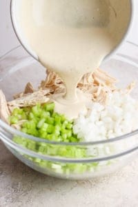 A creamy sauce is being poured into a bowl containing shredded chicken, diced onions, and chopped celery.