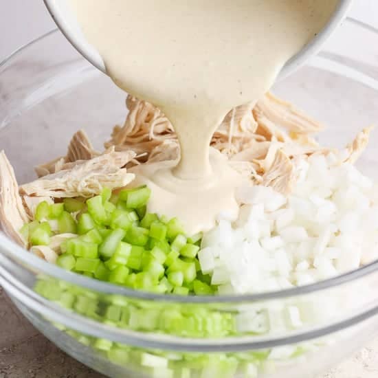 A creamy sauce is being poured into a bowl containing shredded chicken, diced onions, and chopped celery.