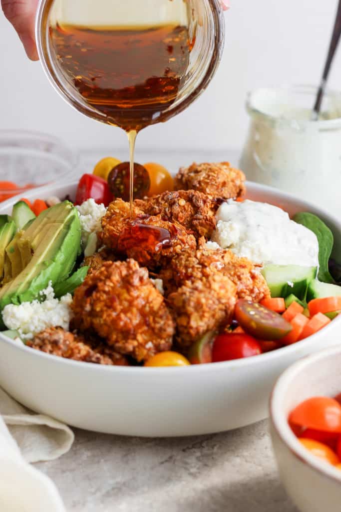 Salad dressing being poured over a bowl of salad with crispy chicken, avocado, tomatoes, and greens.