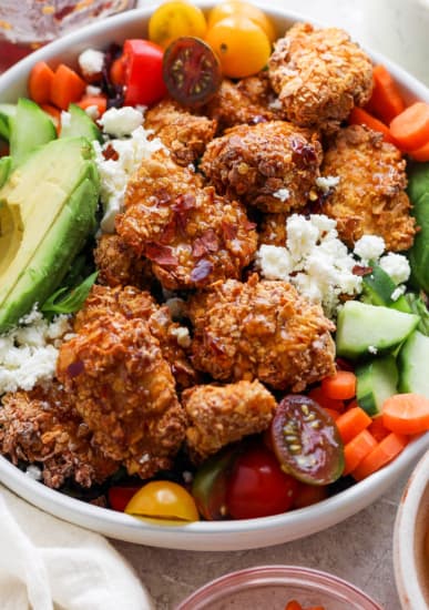 A bowl filled with crispy fried chicken, avocado slices, cherry tomatoes, cucumbers, carrots, and crumbled cheese, presenting a colorful and appetizing mixed meal.