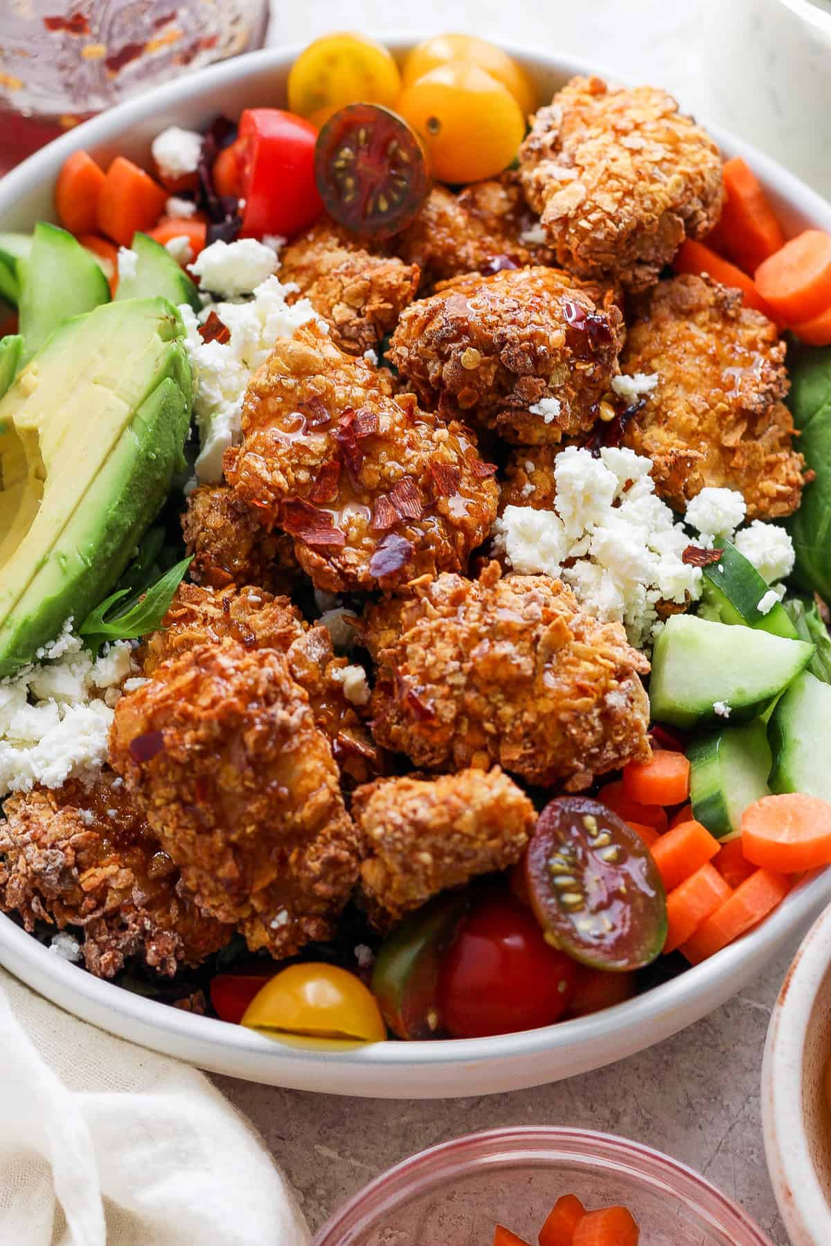 A bowl filled with crispy fried chicken, avocado slices, cherry tomatoes, cucumbers, carrots, and crumbled cheese, presenting a colorful and appetizing mixed meal.