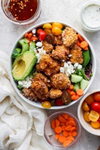 A colorful salad bowl with quinoa, mixed greens, avocado slices, cherry tomatoes, cucumbers, carrots, and topped with crumbled cheese and breaded chicken pieces, accompanied by two small bowls.