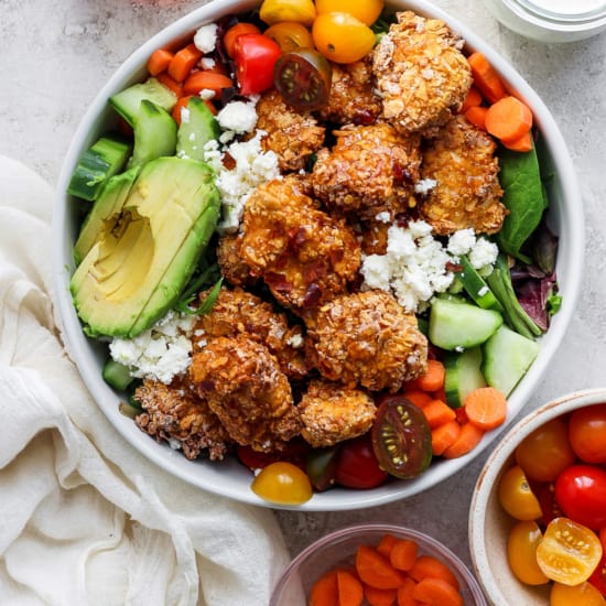 A colorful salad bowl with quinoa, mixed greens, avocado slices, cherry tomatoes, cucumbers, carrots, and topped with crumbled cheese and breaded chicken pieces, accompanied by two small bowls.