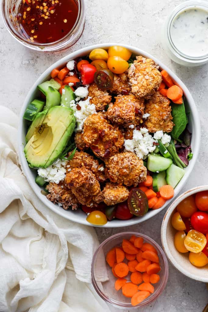 A colorful salad bowl with quinoa, mixed greens, avocado slices, cherry tomatoes, cucumbers, carrots, and topped with crumbled cheese and breaded chicken pieces, accompanied by two small bowls.