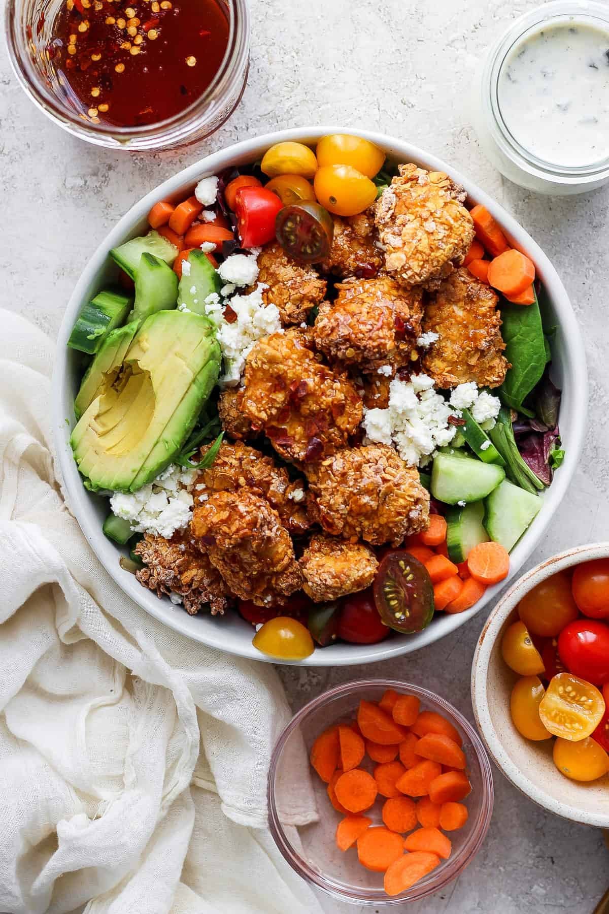 A colorful salad bowl with quinoa, mixed greens, avocado slices, cherry tomatoes, cucumbers, carrots, and topped with crumbled cheese and breaded chicken pieces, accompanied by two small bowls.