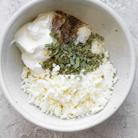 A bowl of crumbled feta cheese mixed with seasonings and yogurt on a textured surface.