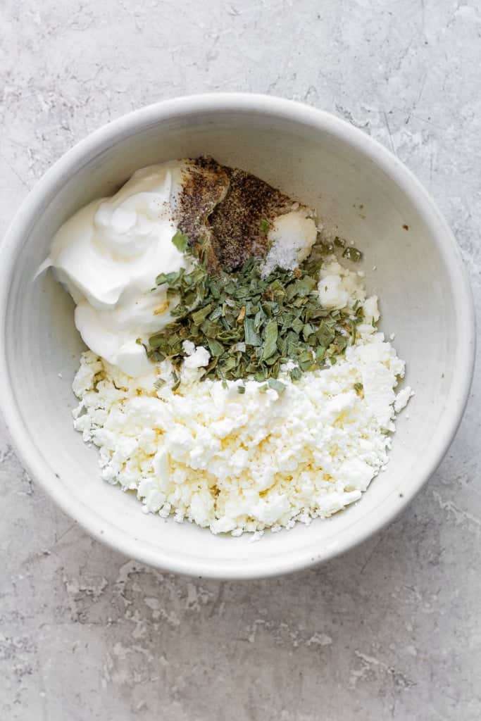 A bowl of crumbled feta cheese mixed with seasonings and yogurt on a textured surface.