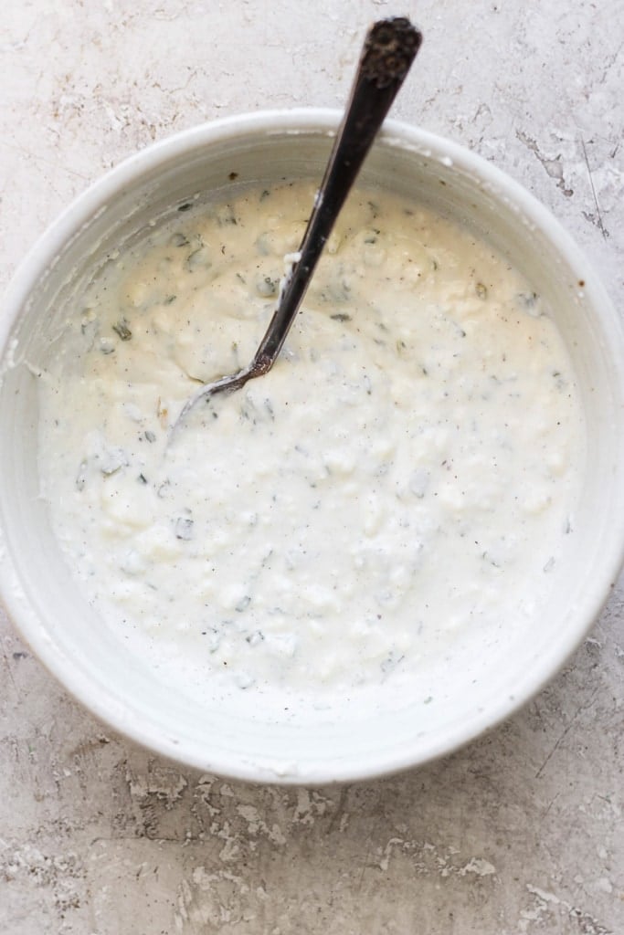 A bowl of creamy herb dressing with a spoon on a textured surface.